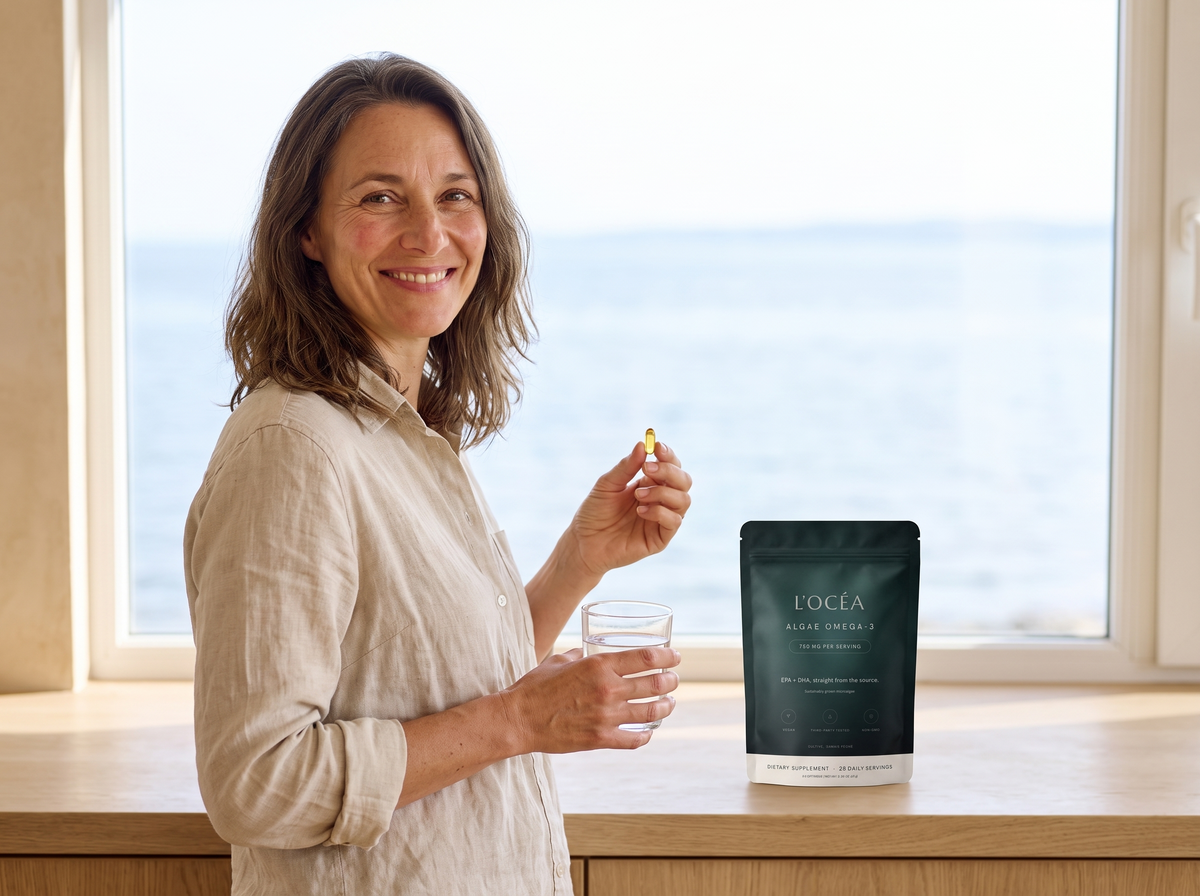 A woman at a bright coastal kitchen counter holding a gold softgel and a glass of water, smiling, the L'Océa pouch on the counter beside her and calm water beyond the window.